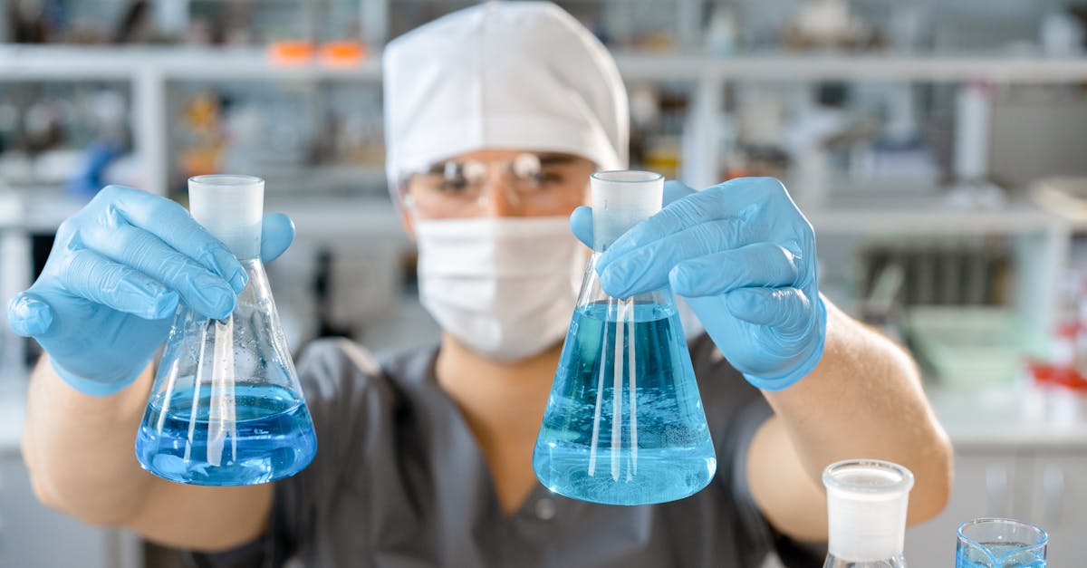 A scientist in protective gear handling blue liquids in laboratory flasks.